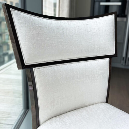 The Classic Deco Style Dining Chair features a glossy dark wood frame and white textured fabric upholstery, positioned near a window with a cityscape view in the background.