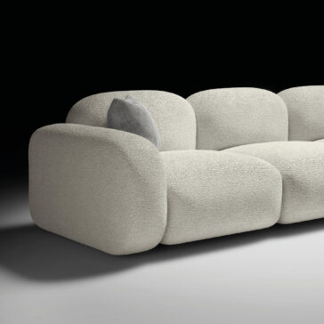 Contemporary Italian Sofa The Contemporary Italian Sofa features a modern, plush design in light beige with rounded edges and soft, textured fabric. It includes a light grey throw cushion. Set against a black background with a grey floor.