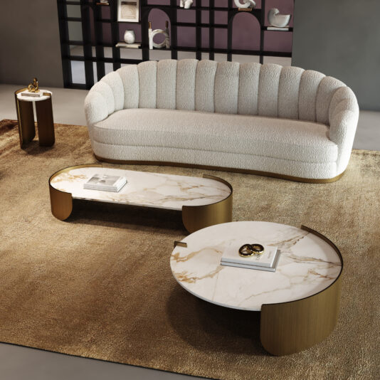 A modern living room with a curved white sofa, contemporary marble coffee tables featuring gold accents, a tan rug, and a round side table with a decorative object. Decor-filled shelves can be seen in the background.