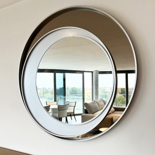 The Contemporary Italian Designer Offset Circles Mirror (Copy) showcases a large and a smaller overlapping circular mirror, reflecting a modern living room with a table, chairs, sofa, and expansive windows overlooking trees and a river.