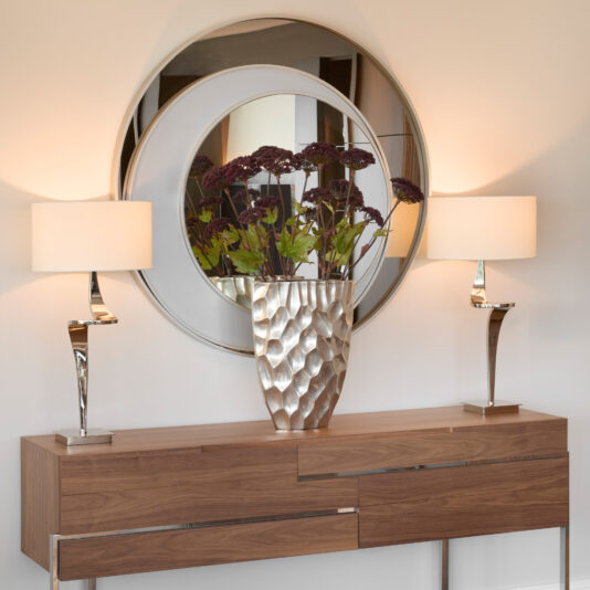 Contemporary Italian Designer Offset Circles Mirror A modern wooden console table with a textured silver vase and two silver lamps sits beneath a Contemporary Italian Designer Offset Circles Mirror (Copy), which reflects the stylish décor arrangement.