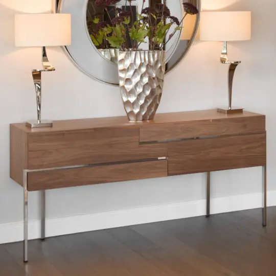 The Contemporary Walnut Veneer Sideboard with metal legs is styled with a large metallic vase of green and burgundy plants, two tall white-shaded lamps, and a round mirror hanging above.