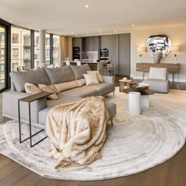 Contemporary Walnut Veneer Sideboard Modern living room with large grey corner sofa, tan fur throw, round coffee tables, circular rug, and open-plan kitchen. The Contemporary Walnut Veneer Sideboard enhances elegance as floor-to-ceiling windows fill the space with light.