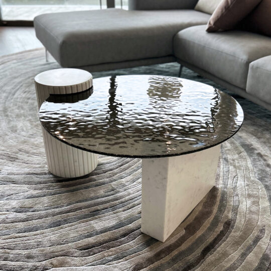 A modern living room showcases the Exclusive Ammonite Tuscany Quartz Coffee Table with a marble base, alongside a small cylindrical side table and grey sofa, atop a patterned circular rug.