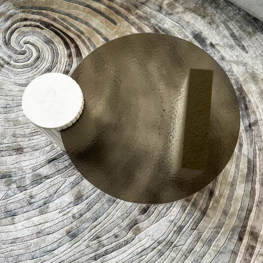 Exclusive Ammonite Tuscany Quartz Coffee Table An Exclusive Ammonite Tuscany Quartz Coffee Table with a textured metallic finish topped by a cylindrical white accent sits on a neutral-toned carpet featuring concentric swirl patterns.
