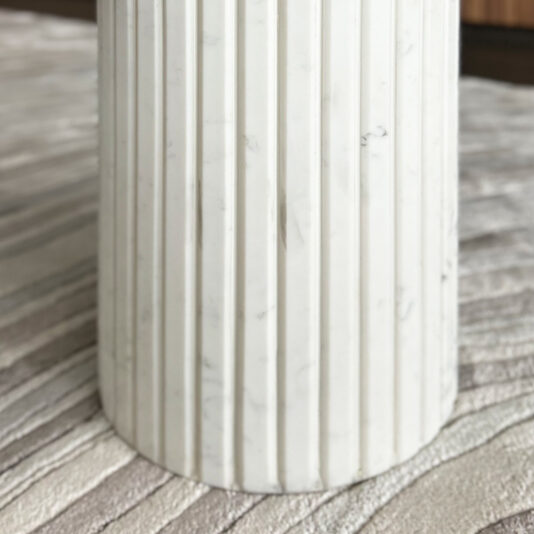 Exclusive Ammonite Tuscany Quartz Coffee Table A close-up of a cylindrical white marble column with vertical grooves stands on a textured, striped surface beside the Exclusive Ammonite Tuscany Quartz Coffee Table.