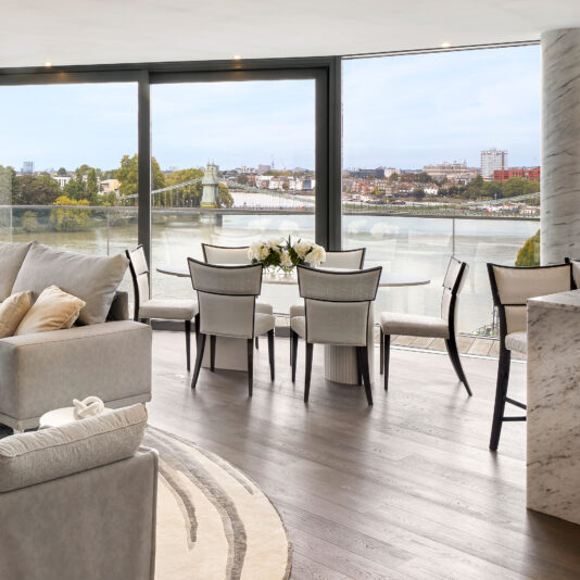 Exclusive Oval Ammonite Tuscany Quartz Dining Set Modern living and dining area with light-coloured furniture, large windows, and a river view. The Exclusive Oval Ammonite Tuscany Quartz Dining Set is featured, accented by a vase of white flowers on the table.