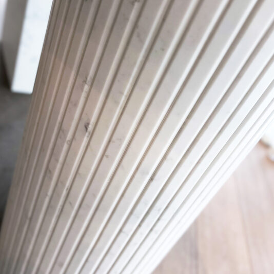 Exclusive Oval Ammonite Tuscany Quartz Dining Table A close-up of a fluted white marble column with vertical grooves, capturing the timeless elegance of the Exclusive Oval Ammonite Tuscany Quartz Dining Table, set before a softly blurred grey and brown background.