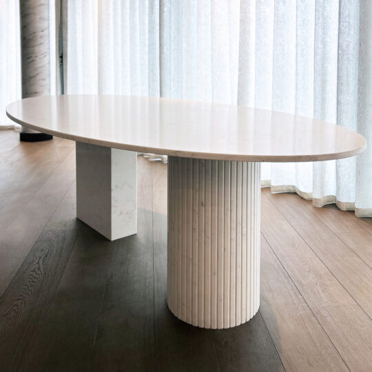 The Exclusive Oval Ammonite Tuscany Quartz Dining Table features a smooth top, supported by a rectangular block and fluted cylindrical base, placed on wooden flooring near large sheer curtains.