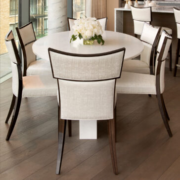 Exclusive Oval Ammonite Tuscany Quartz Dining Table An Exclusive Oval Ammonite Tuscany Quartz Dining Table, adorned with a vase of white flowers, is surrounded by six upholstered chairs with dark wooden legs on a wooden floor beside large windows.