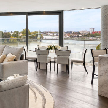 Exclusive Oval Ammonite Tuscany Quartz Dining Table A modern living and dining area with light furniture, floor-to-ceiling windows, an Exclusive Oval Ammonite Tuscany Quartz Dining Table for six, and a balcony overlooking a river and cityscape.