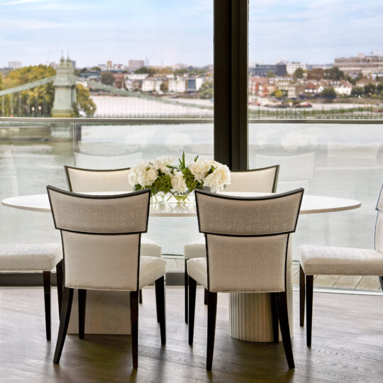 A modern dining area showcases four white chairs around an Exclusive Oval Ammonite Tuscany Quartz Dining Table with a floral centrepiece. Large windows frame scenic river and city views, with a picturesque bridge in the background. in a luxury contemporary London riverside apartment