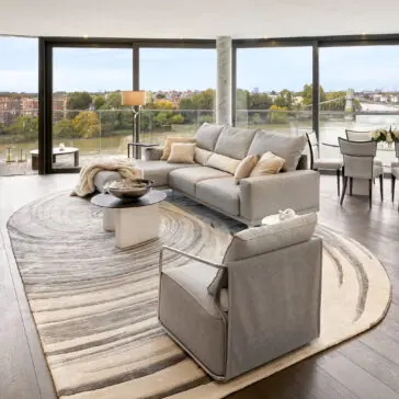 A modern living room with floor-to-ceiling windows overlooks a river and city, featuring a grey sofa, armchair, and the High Quality Hand Woven Swirl Rug. Neutral tones and an open dining area create an airy atmosphere.