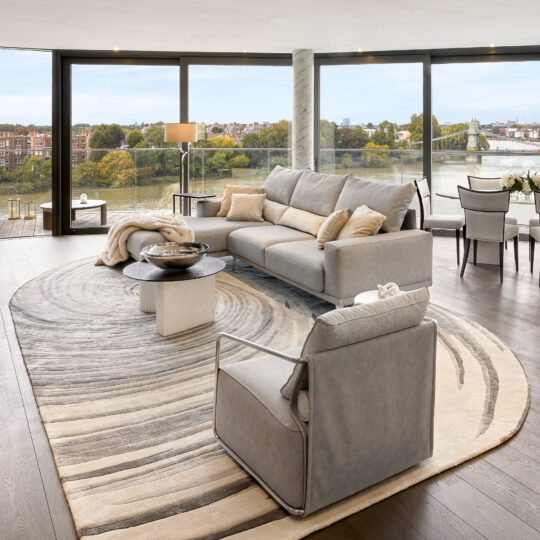 A modern living room with floor-to-ceiling windows overlooks a river and city, featuring a grey sofa, armchair, and the High Quality Hand Woven Swirl Rug. Neutral tones and an open dining area create an airy atmosphere.