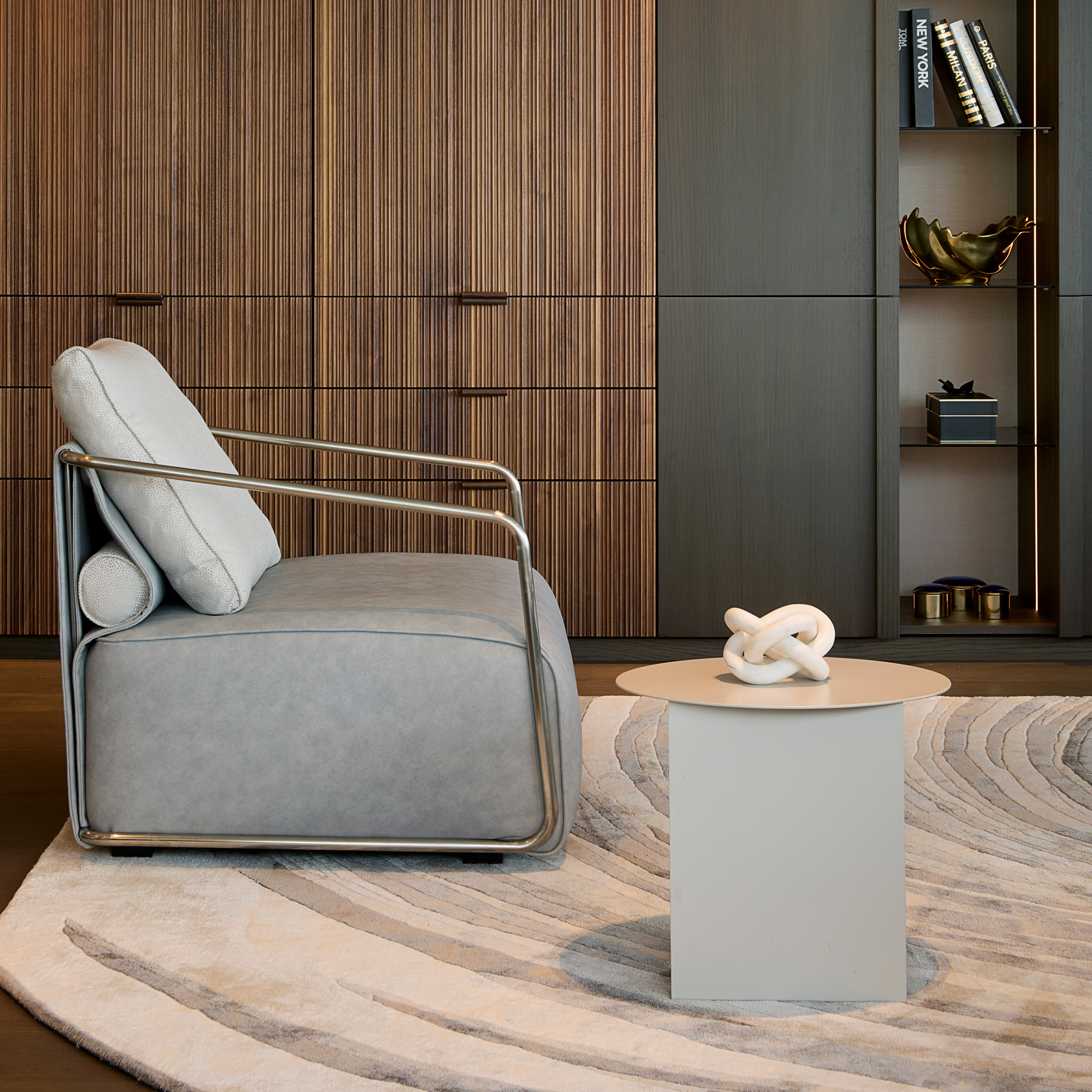 A modern living room with a light grey armchair, round side table topped with a white knot sculpture, wooden cupboards, shelves of books and decor—all anchored by the High Quality Hand Woven Swirl Rug and stylish patterned accents.