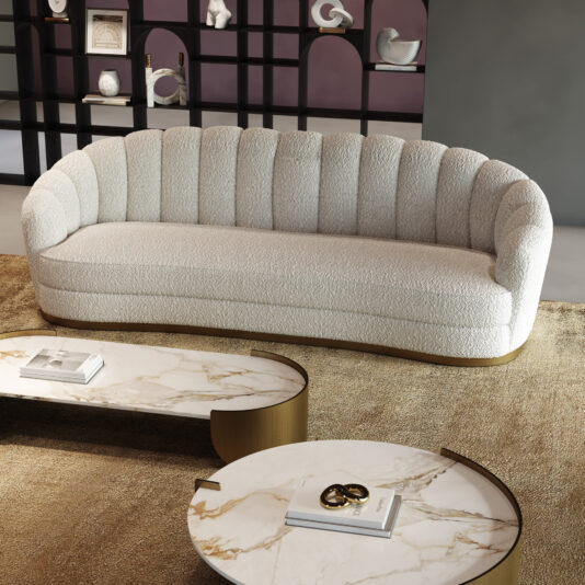 A Large Modern Velvet Sofa in plush cream sits on a beige rug next to two marble-topped coffee tables with gold accents. Black shelves in the background display white decor.