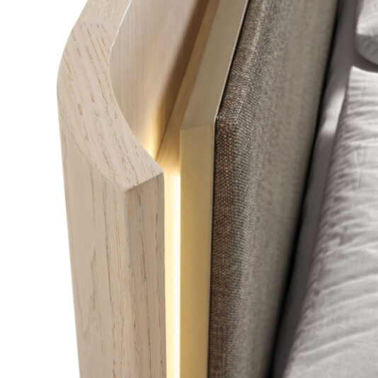 Contemporary Bed With Backlit Headboard Close-up of the Contemporary Bed With Backlit Headboard, featuring smooth, light wood with curved edges, a brown upholstered headboard, and soft grey bedding. Warm lighting glows subtly between the headboard and wood.