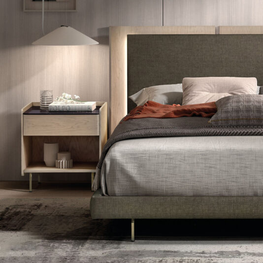 Contemporary Bedside With Porcelain Marble Top A modern bedroom with a grey and brown neatly made bed, a large padded headboard, and a contemporary bedside with porcelain marble top decorated with books, flowers, and a white lamp.