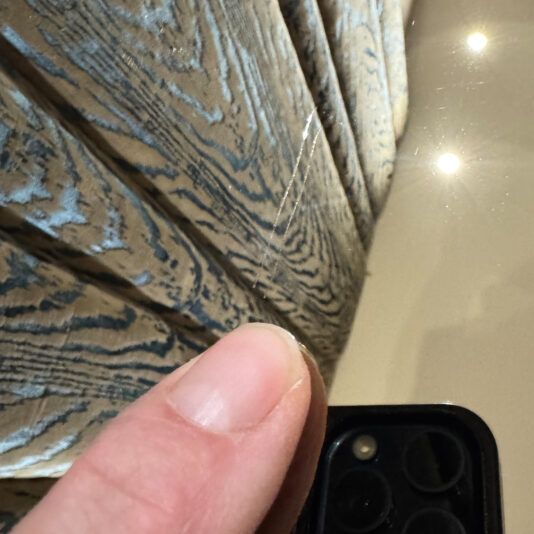 Luxury Italian Designer Seashell Effect 3 Door Buffet A person’s thumb holds a triple-camera phone reflecting beige and blue fabric and ceiling lights, capturing the same elegance and sheen as the Luxury Italian Designer Seashell Effect 3 Door Sideboard.