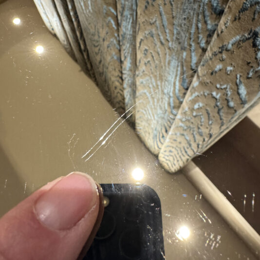 Luxury Italian Designer Seashell Effect 3 Door Buffet A close-up of a person's thumb by the shiny surface of the Luxury Italian Designer Seashell Effect 3 Door Sideboard, showing noticeable scratches and reflected ceiling lights, with patterned fabric or a curtain in the background.