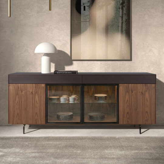 A Modern 4 Door Walnut Veneer Sideboard with wood and glass doors showcases crockery, topped with a white lamp and book, placed against a textured wall beneath abstract artwork.