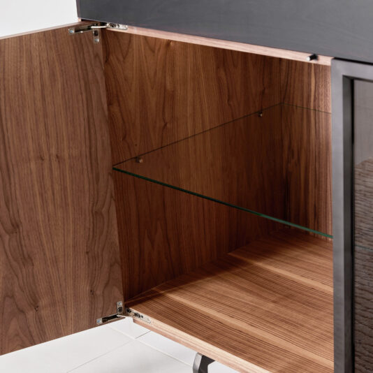 The Modern 4 Door Walnut Veneer Sideboard features an open door that reveals an empty interior with a glass shelf and wood grain finish, all elevated on sleek black metal legs.