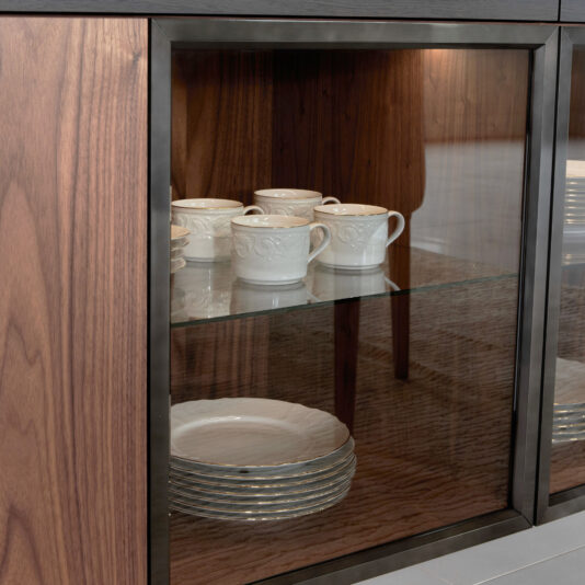The Modern 4 Door Walnut Veneer Sideboard features a glass-fronted cabinet displaying eight white teacups with saucers on the upper shelf and two stacks of decorative-edged plates below, all set against a stylish wood interior.