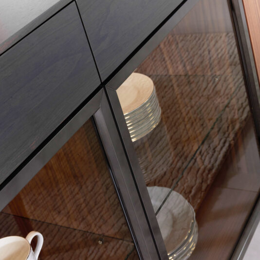Close-up of the Modern 4 Door Walnut Veneer Sideboard featuring a dark-framed glass front, stacked white plates on glass shelves, a white cup, and a wood-textured back panel.