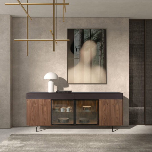 A modern space showcases the Modern 4 Door Walnut Veneer Sideboard with glass doors displaying crockery, topped with a white table lamp and book. Overhead, a geometric gold chandelier hangs beside abstract art on a textured beige wall.
