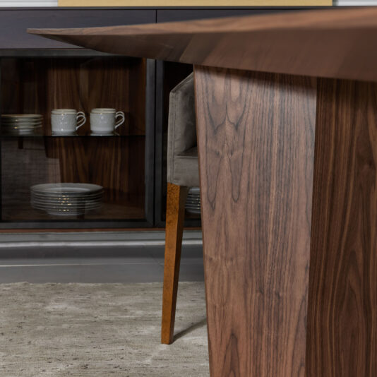 Modern Rectangular Veneer Dining Table Close-up of the Modern Rectangular Veneer Dining Table showing its grain, beside a grey upholstered chair with wooden legs. In the background, a cupboard displays stacked plates and cups behind glass doors.