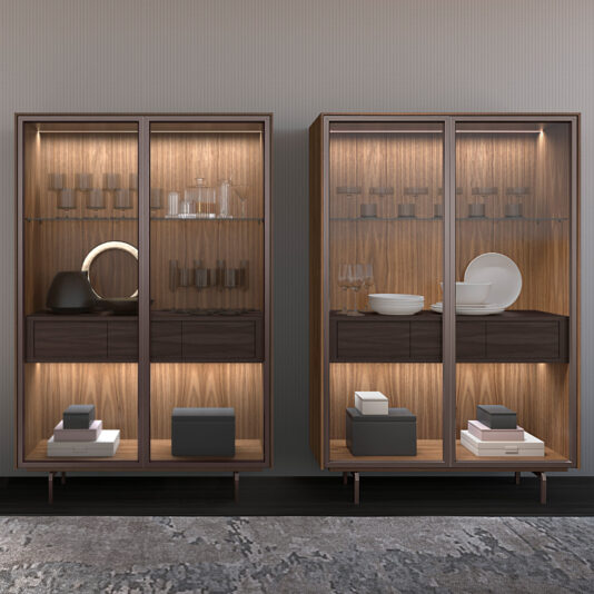 Two Modern Walnut Veneer Display Cabinets (Copy) with glass doors display glassware, dishes, and décor. Interior lighting highlights the items, boxes sit on bottom shelves, and a patterned rug lies in front.
