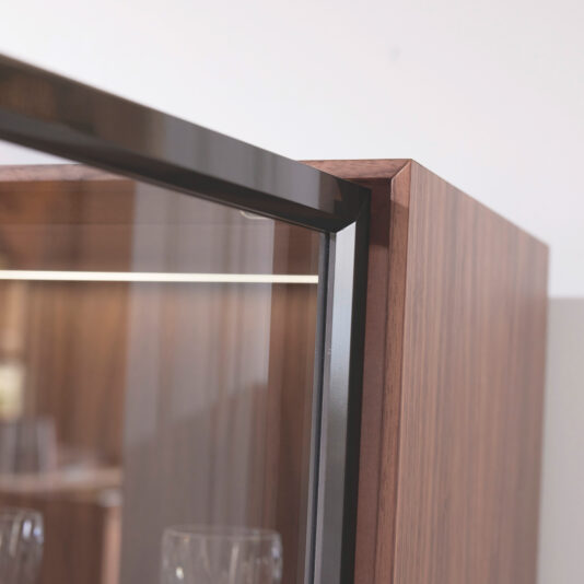 Modern Walnut Veneer Display Cabinet Close-up of the Modern Walnut Veneer Display Cabinet’s top corner, highlighting its dark wood frame, reflective glass door, metal trim, and softly lit blurred glassware inside against a neutral background.