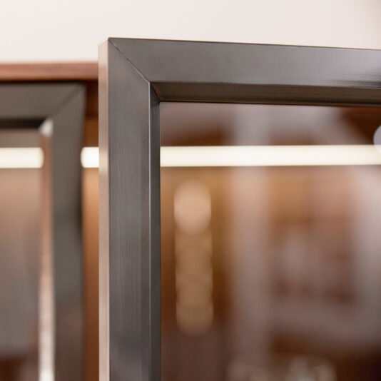 Modern Walnut Veneer Display Cabinet Close-up of the corner of a glass door with a sleek metal frame, set against a soft brown and beige background—perfect for highlighting the elegant design details of the Modern Walnut Veneer Display Cabinet.