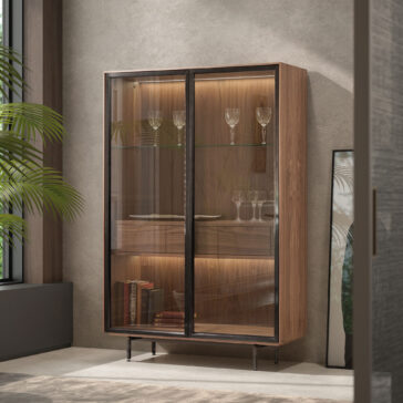 Modern Walnut Veneer Display Cabinet The Modern Walnut Veneer Display Cabinet with glass doors stands against a textured wall, its warm interior lighting illuminating displayed glassware. A nearby leafy plant adds a fresh touch of greenery to the stylish scene.