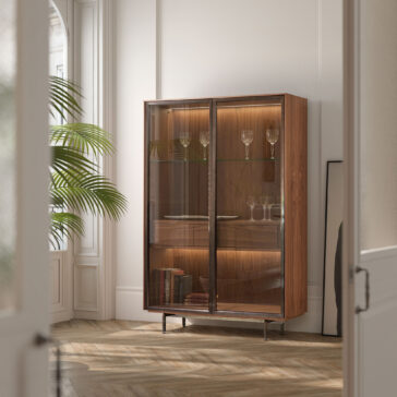Modern Walnut Veneer Display Cabinet The Modern Walnut Veneer Display Cabinet, featuring glass doors, displays glassware and books. It stands against a light wall in a bright room with herringbone wood floors and a large green plant nearby.