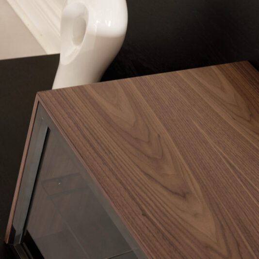Modern Walnut Veneer TV Cabinet A close-up of the Modern Walnut Veneer TV Cabinet’s wooden surface with a glass panel edge, set beside a white abstract ceramic sculpture against a dark background.