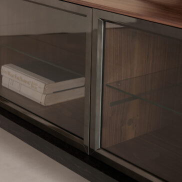 A close-up of the Modern Walnut Veneer TV Cabinet shows its sleek, modern design and rich dark wood finish. Glass doors reveal stacked books on the left shelf, topped by a glass shelf inside.