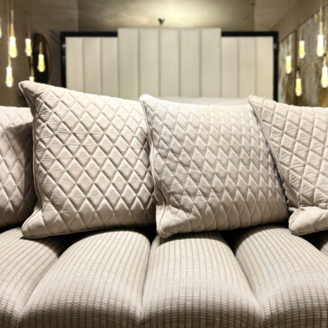 A Set Of 4 Quilted Nubuck Scatter Cushions in beige sits on a textured, light sofa, softly lit against a modern minimalistic backdrop with vertical panels and pendant lights.