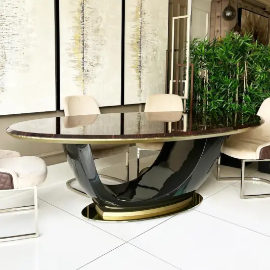 A modern dining room features a glossy oval table with a curved black and gold base, surrounded by beige upholstered chairs with metal frames, white tiled floors, wall art, and tall green plants in the background.