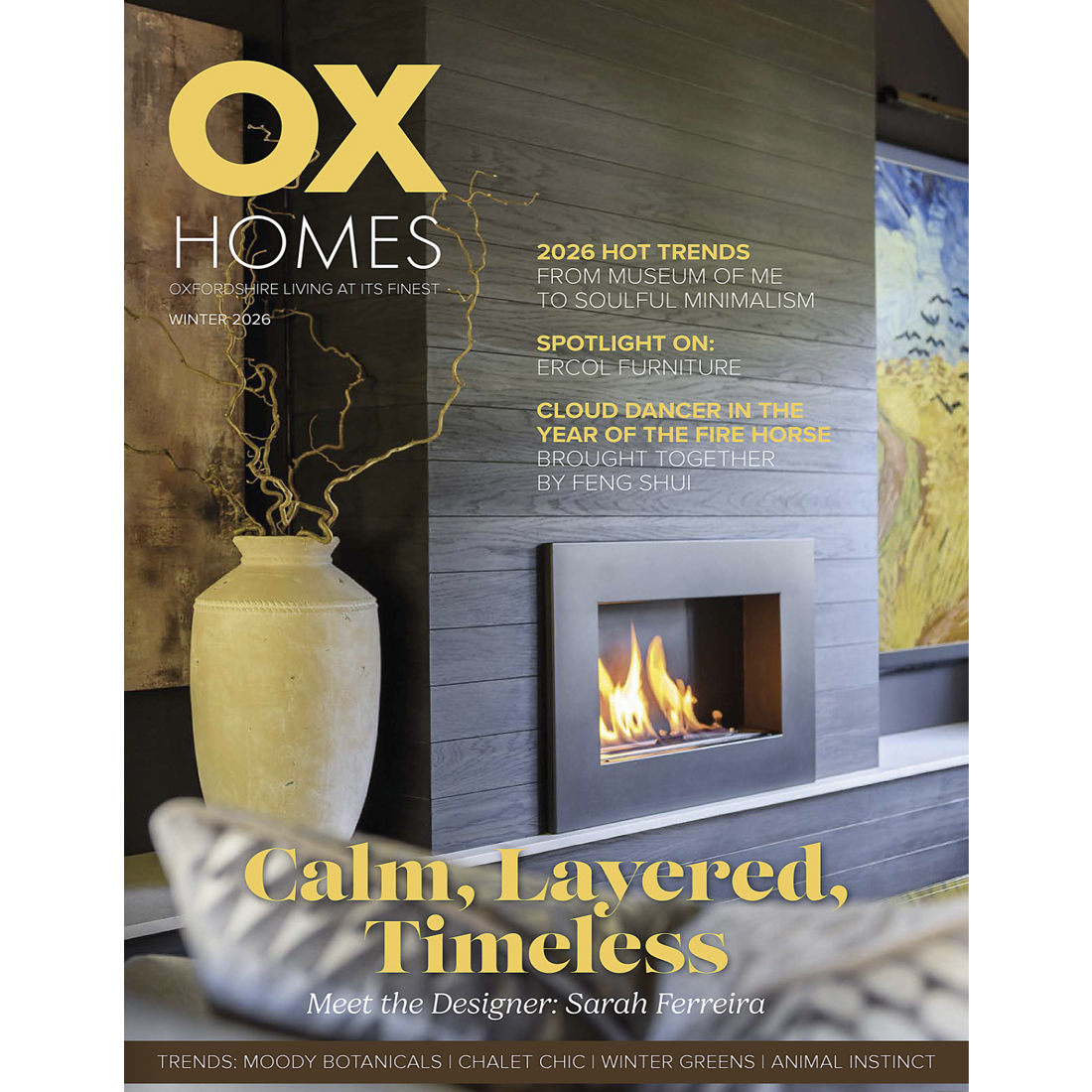 A modern living room with a built-in fireplace, stacked firewood, and decorative accents. The magazine cover features headlines about design trends and the quotation "Calm, Layered, Timeless" by designer Sarah Ferreira.