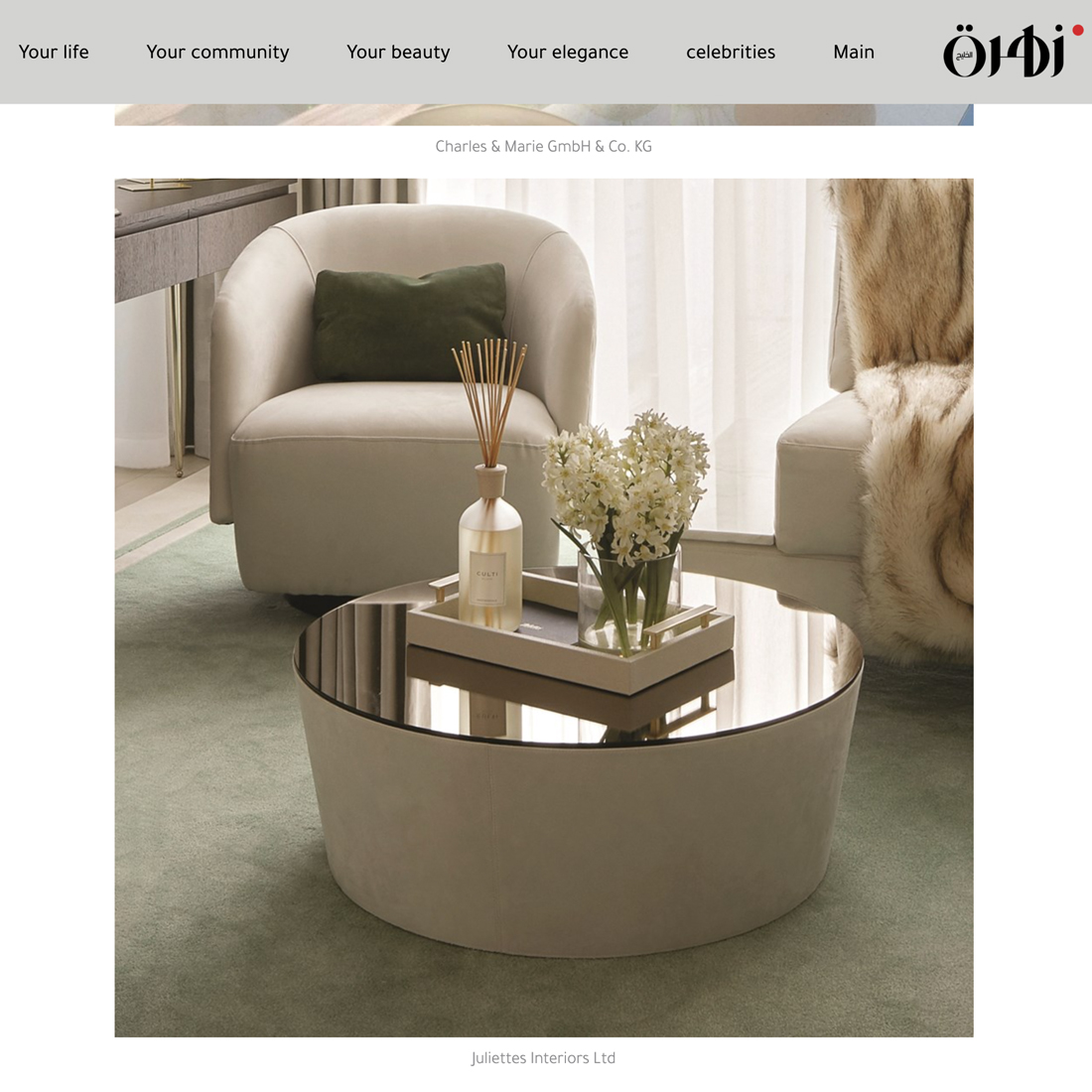 A modern living room with a round, mirrored coffee table holding a tray, white flowers, and a reed diffuser. An armchair with a green cushion sits nearby on a light green rug. Light flows through sheer curtains.