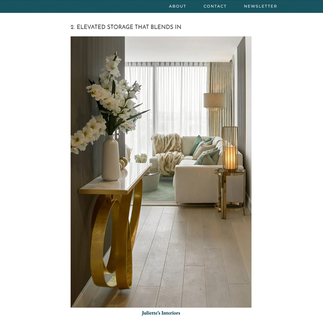 A modern living space with a white and gold console table holding white flowers, light wood floors, a green sofa with cushions and throws, a round side table, and sheer curtains letting in natural light.
