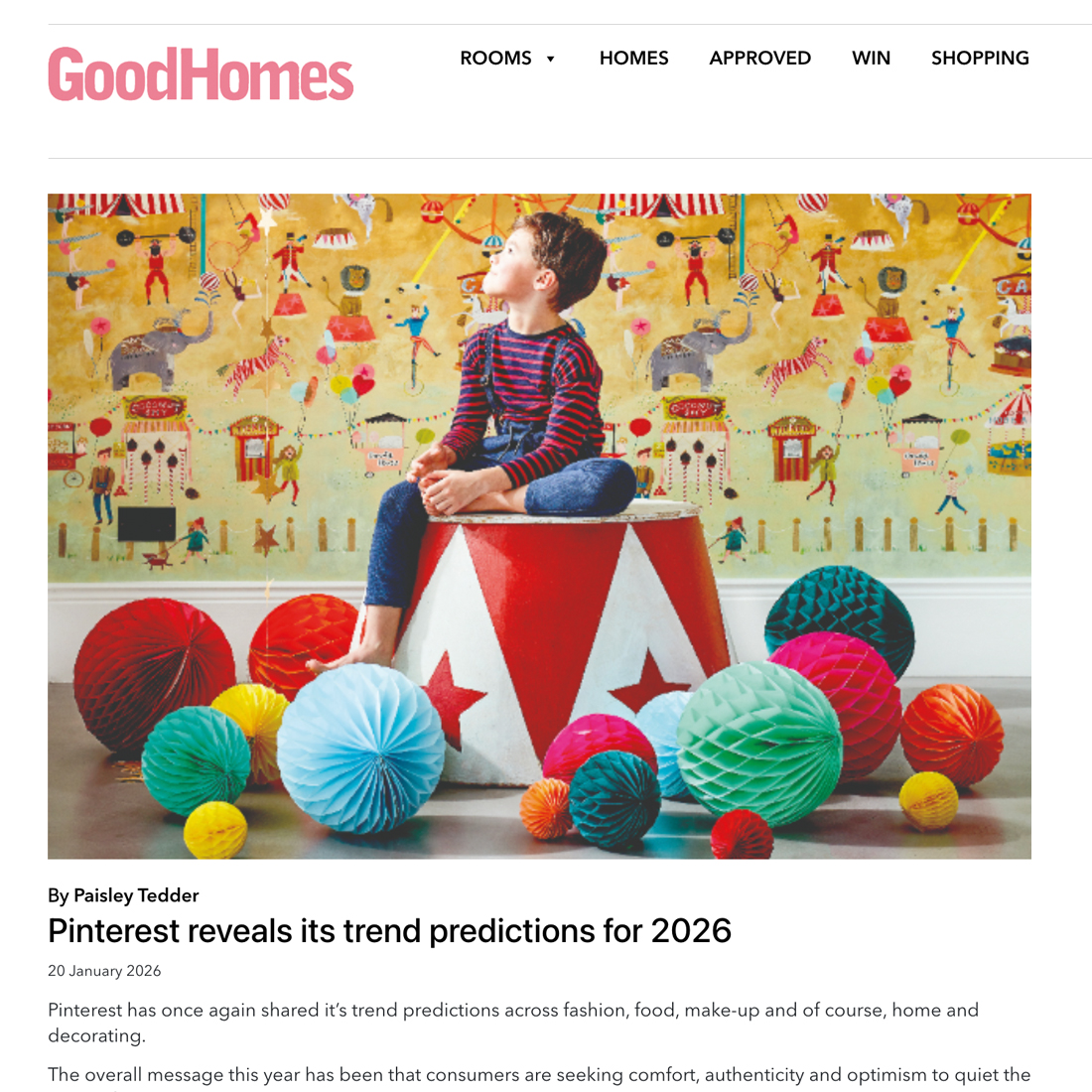 A young boy in a colourful jumper and jeans sits on a large red and white circus drum, surrounded by vibrant honeycomb paper decorations. He looks sideways, with vintage-style wallpaper behind him.