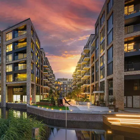 Modern apartment blocks with large windows and balconies line a narrow waterway at sunset, reflecting warm light in the water. The sky is vibrant with orange and purple clouds. Some lights are on inside the buildings.