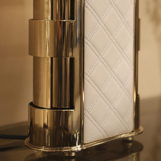 Luxury Hand-Stitched Leather Table Lamp Close-up of the Luxury Quilted Faux Leather Table Lamp base, featuring a diamond-patterned quilted white leather panel and a shiny gold-tone metallic finish standing on four small feet.