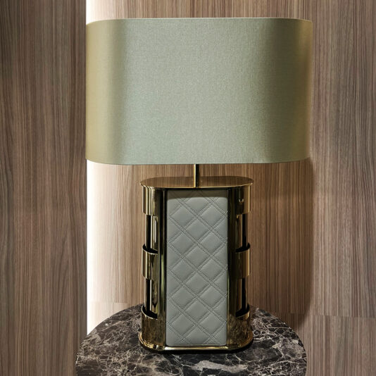 The Luxury Quilted Faux Leather Table Lamp, featuring a rectangular base and a large cylindrical light grey shade, rests on a black marble surface in front of a wooden backdrop.