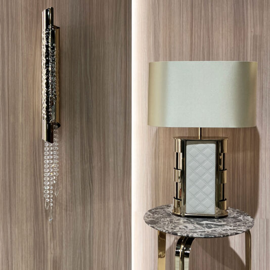 Luxury Hand-Stitched Leather Table Lamp The Luxury Quilted Faux Leather Table Lamp with a white base is placed on a round marble-topped side table, whilst a wall-mounted metallic fitting with hanging crystal beads highlights the wood-textured backdrop nearby.