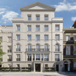 A grand, multi-storey light stone building with tall windows, ornate railings, and a symmetrical facade, set on a cobbled street under a blue sky with scattered clouds.