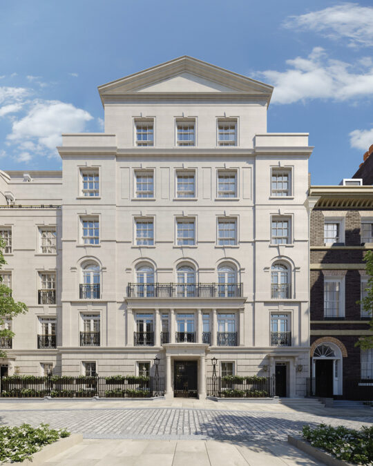 A grand, multi-storey light stone building with tall windows, ornate railings, and a symmetrical facade, set on a cobbled street under a blue sky with scattered clouds.