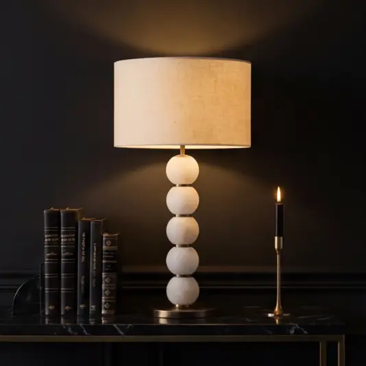 The 5-Globe Tiered Table Lamp, featuring a beige shade and stacked white sphere base, is displayed on a dark marble table beside a brass candlestick with a black candle and gold-accented books against a dark wall.